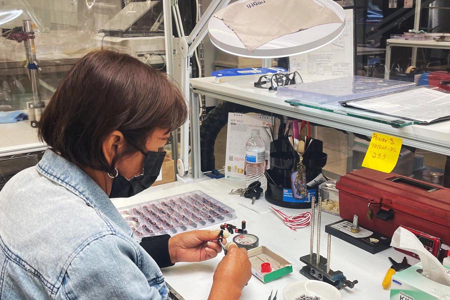 Photograph of a female worker assembling sensors at Magnetic Sensors Corporation using US-made components. The worker is seated at a workbench with various tools and components around her. She is wearing grounding bracelet to protect her from electrical voltage,.